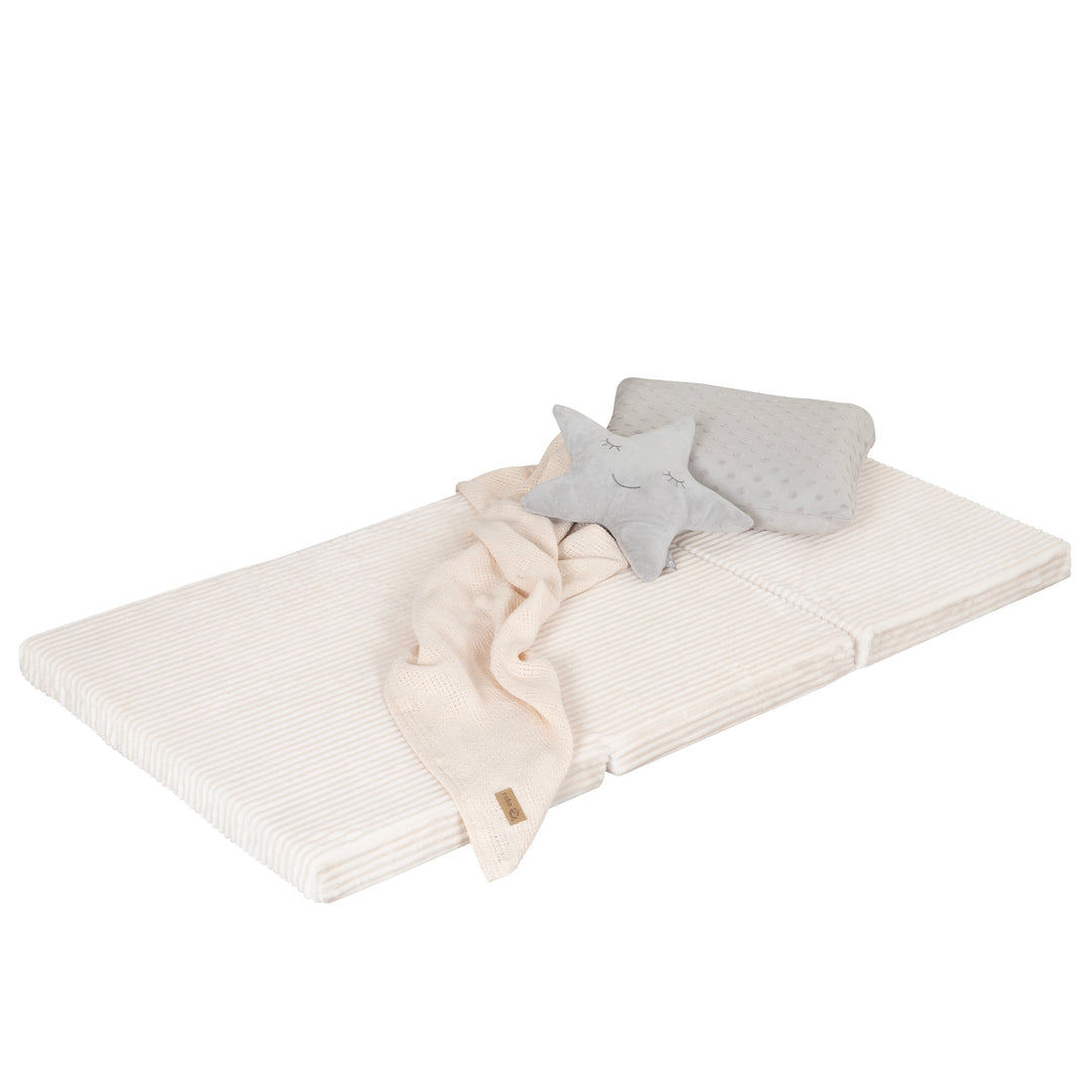 Freisteller der ausgeklappten Reisebettmatratze in Beige mit Cord-Bezug, dekoriert mit Plüsch-Stern, grau gepunktetem Kissen und cremefarbener Strickdecke, vor weißem Hintergrund.