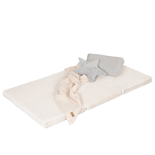 Freisteller der ausgeklappten Reisebettmatratze in Beige mit Cord-Bezug, dekoriert mit Plüsch-Stern, grau gepunktetem Kissen und cremefarbener Strickdecke, vor weißem Hintergrund.