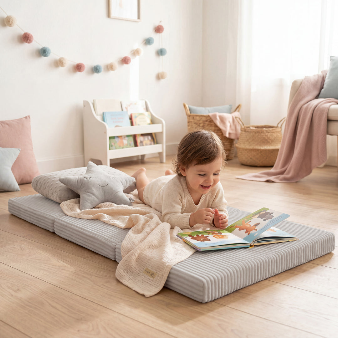 Lifestyle-Bild: Kleinkind in cremefarbenem Strickpulli liegt bäuchlings auf der ausgeklappten Reisebettmatratze in Grau und liest ein Bilderbuch, im Hintergrund ein Zimmer mit Bücherregal, Bommel-Girlande und rosafarbenen Kissen.
