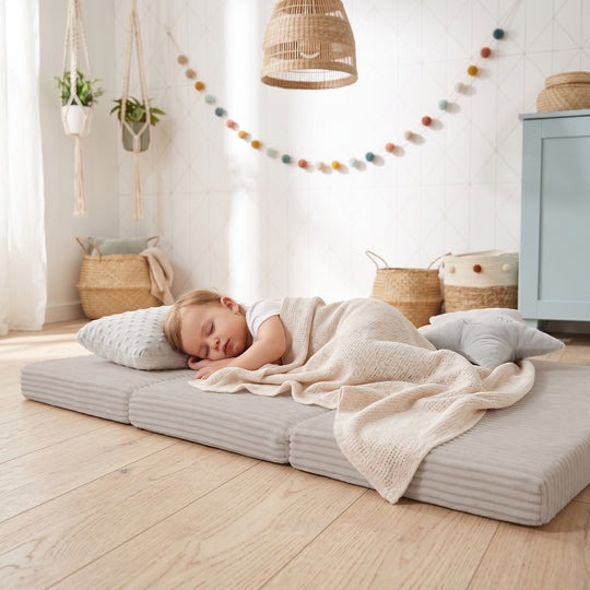 Lifestyle-Bild: Schlafendes Kleinkind, in eine gestrickte Decke gehüllt, liegt auf der ausgeklappten Reisebettmatratze in Grau neben einem Plüsch-Stern, im Hintergrund ein Kinderzimmer mit Makramee-Pflanzenampeln, Bommel-Girlande und Rattan-Hängelampe.