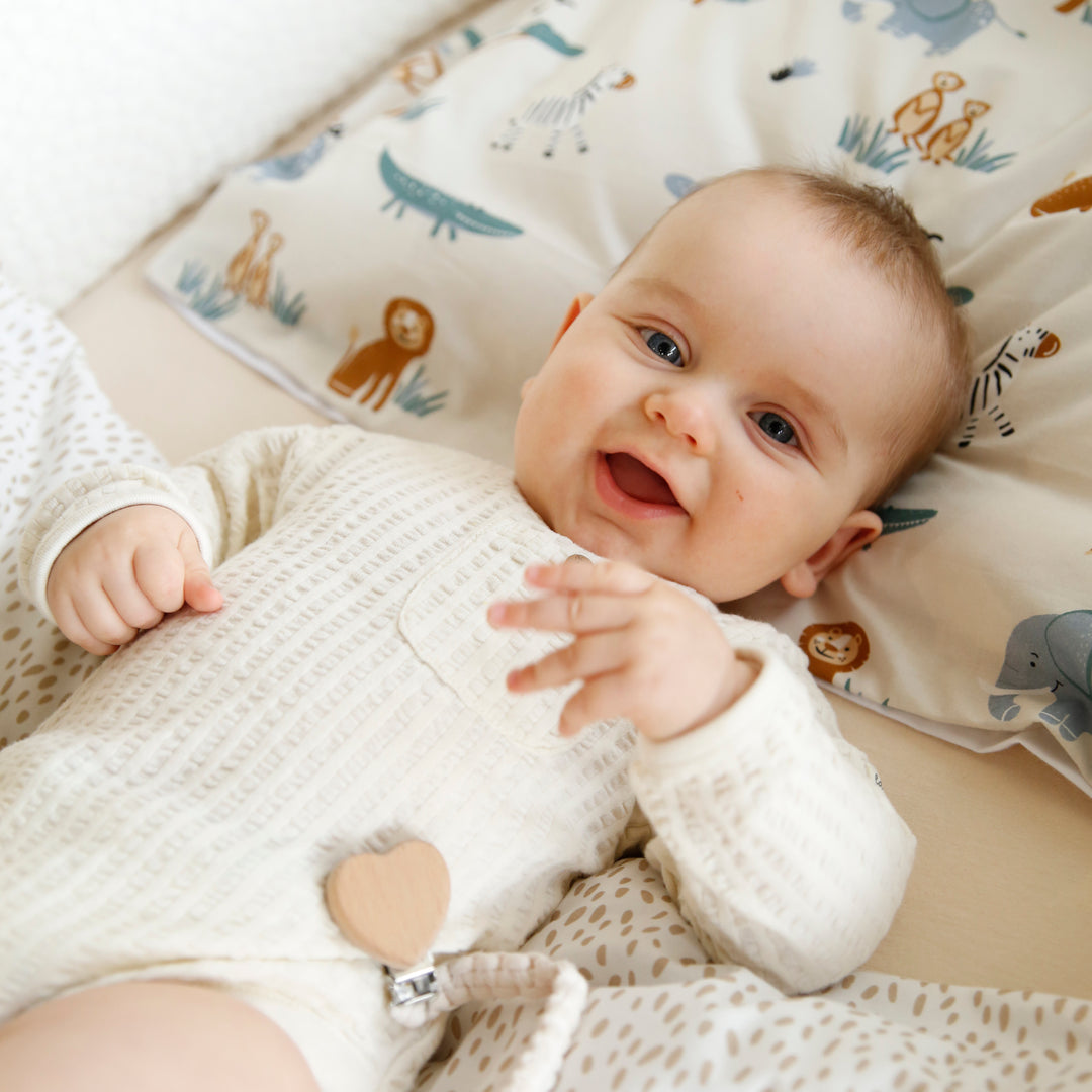 Lachendes Baby im gestrickten Strampler liegt auf der Wiegenbettwäsche "Savanna Friends" mit Tiermotiven im Hintergrund