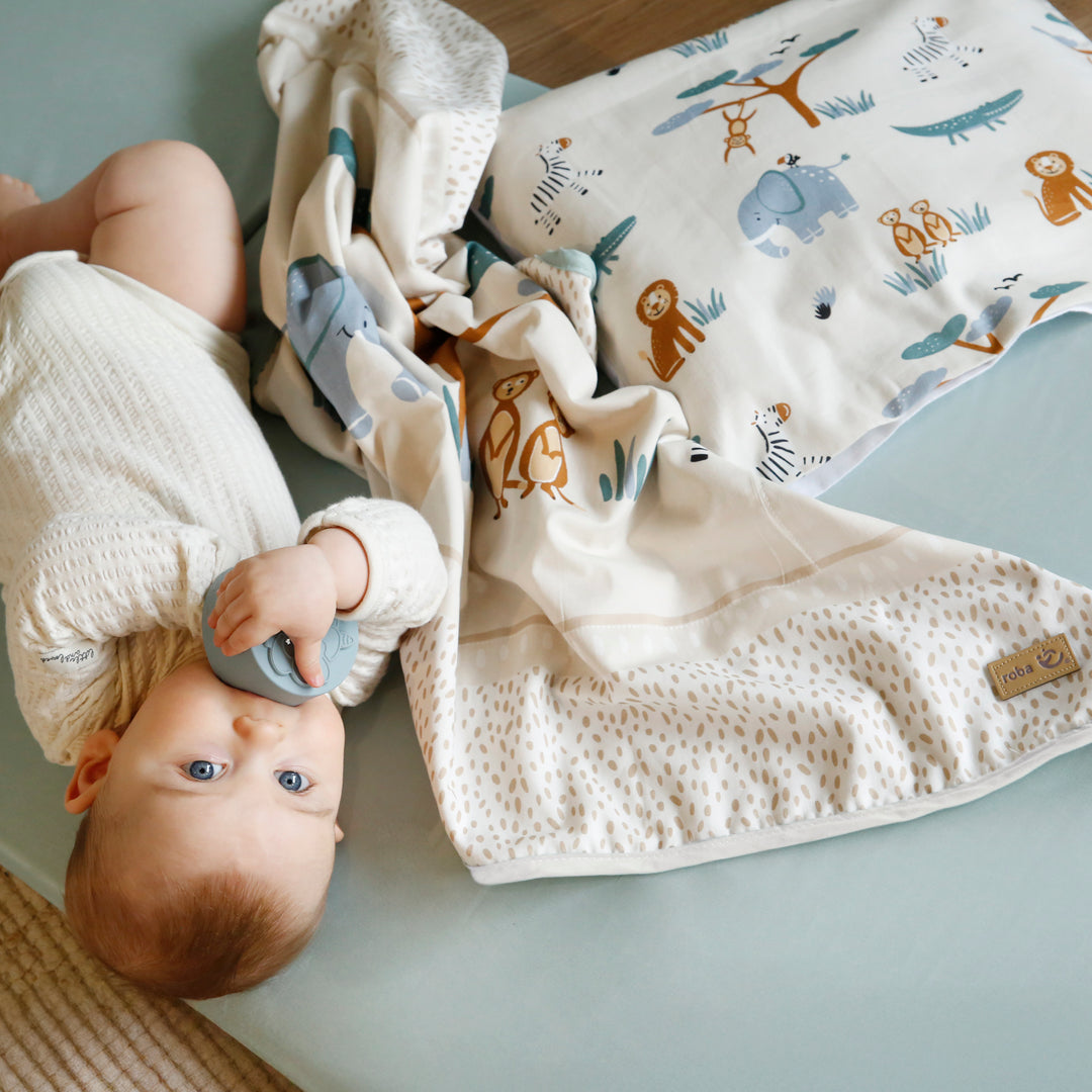 Baby liegt kuschelnd neben der Babydecke "Savanna Friends" mit buntem Tiermotiv – Elefant, Löwe, Zebra und Affe