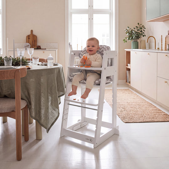 Vergleichsbild: links ein Baby im weißen Lernturm in Hochstuhl-Konfiguration mit Sternkissen, rechts ein lachendes Kleinkind, das in der Steh-Konfiguration steht; am Lernturm hängt ein Anhänger mit der Aufschrift 'SIT & STEP'.
