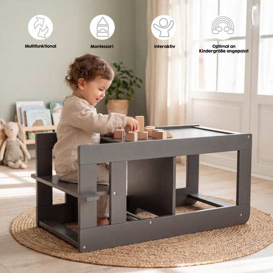 Lifestyle-Bild mit Symbolen 'Multifunktional, Montessori, Interaktiv, Optimal an Kindergröße angepasst'; Kleinkind sitzt an der umgebauten Sitzgarnitur des Anthrazit-Lernturms und spielt mit Holzbauklötzen in einem Wohnzimmer mit Bücherregal, Kuscheltier und Jute-Teppich.