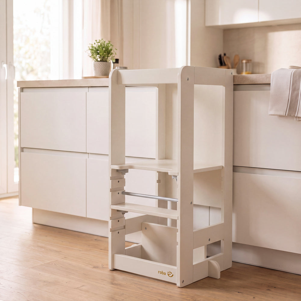 Lernturm 'Step & Play' in Cashmere, leer in einer hellen Küche neben einer weißen Kochinsel mit Geschirrtuch und Grünpflanze auf der Arbeitsplatte.