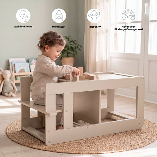 Lifestyle-Bild mit Symbolen 'Multifunktional, Montessori, Interaktiv, Optimal an Kindergröße angepasst'; Kleinkind sitzt an der umgebauten Sitzgarnitur des Cashmere-Lernturms und spielt mit Holzbauklötzen in einem Wohnzimmer mit Bücherregal, Kuscheltier und Jute-Teppich.