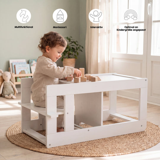 Lifestyle-Bild mit Symbolen 'Multifunktional, Montessori, Interaktiv, Optimal an Kindergröße angepasst'; Kleinkind sitzt an der umgebauten Sitzgarnitur des weißen Lernturms und spielt mit Holzbauklötzen in einem Wohnzimmer mit Bücherregal, Kuscheltier und Jute-Teppich.
